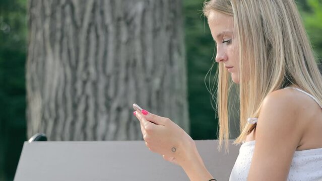 A young Caucasian girl is typing a message on a smartphone while sitting on a bench in the summer. Side view of a girl typing text on a phone in the park.