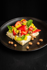 Top view of fruit tart with strawberries, mango, mint and kiwi, on a black plate, black table, vertical, with copy space