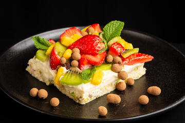 Close-up of fruit tart with strawberries, mango, mint, kiwi and chocolate balls on black plate, black table, horizontal