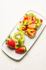 Aerial view of fruit tart with strawberries, mango, mint, kiwi and chocolate balls on a white plate, white table, vertical, with copy space