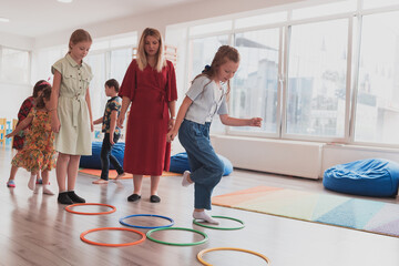 Fototapeta premium Small nursery school children with female teacher on floor indoors in classroom, doing exercise. Jumping over hula hoop circles track on the floor.