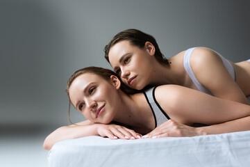 Obraz premium smiling lesbian woman looking away while relaxing on bed near brunette girlfriend on grey background.