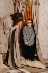 A little boy with Down Syndrome in a shirt and glasses plays hide and seek with his mother. Mom and...