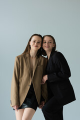 young and happy lesbian partners in blazers holding hands isolated on grey.
