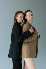 young and tender woman hugging lesbian girlfriend standing with closed eyes isolated on grey.