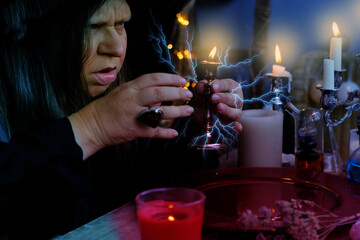female witch holds candle in candlestick in her hands, conjures in dark room, magic items on table,...