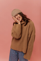 Cute young caucasian girl is smiling looking into camera with half of face covered by hand on pink background. Brunette with wavy hair is wearing hat, sweater and jeans. Concept of enjoying moments