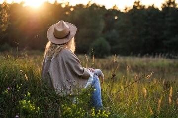 Relaxation outdoors at sunset. Boho woman with hat and poncho sitting in meadow. Enjoyment of...