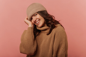 Vivacious young brunette caucasian woman is smiling looking at camera on pink background. Model wears brown hat and sweater. Lifestyle concept sincere emotions of people.