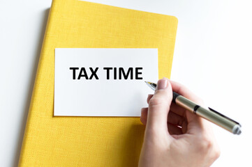 Tax Time inscription on the card on the background of blonde on a bright table next to the hand of a person with a pen writes text