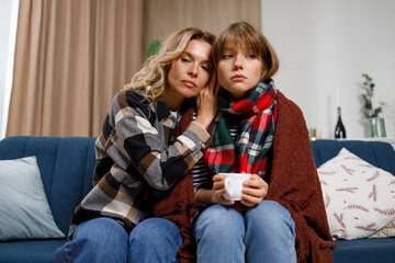 Sad girl with flu in a blanket and with a cup of tea and her mother sitting on the sofa in the living room. Pretty mature woman hugged her beloved daughter who caught a cold