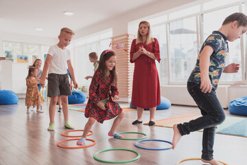 Small nursery school children with female teacher on floor indoors in classroom, doing exercise. Jumping over hula hoop circles track on the floor.