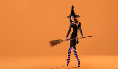 A ballerina on pointe shoes in a black witch costume in a hat and with a broom dances on an orange...