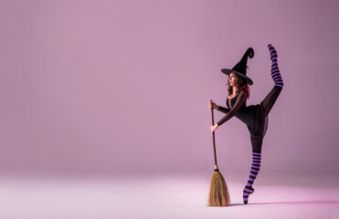 ballerina on pointe shoes in a black witch costume in a hat and with a broom is dancing on the roof on a lilac background. © Maria Moroz