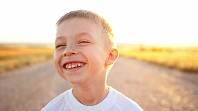 Close up portrait and face of caucasian baby boy. Child stands on asphalt road next to wheat fields. Kid happy smiling and toothy laughs on walk in sun rays. Nature landscape. Leisure activity. Rest.