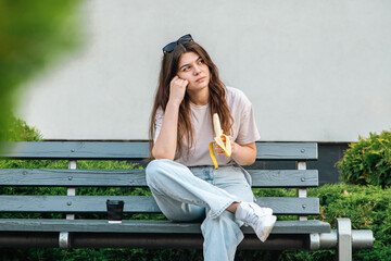 A sad young woman sits on a bench and eats a banana.