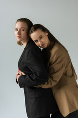 brunette woman hugging lesbian partner in black blazer and looking at camera isolated on grey.