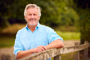 Portrait Of Smiling Casually Dressed Mature Or Senior Man Leaning On Fence On Walk In Countryside
