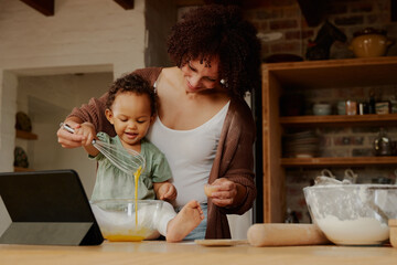Happy biracial mother and daughter whisking egg near digital tablet in kitchen at home