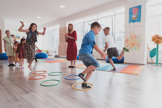 Small nursery school children with female teacher on floor indoors in classroom, doing exercise. Jumping over hula hoop circles track on the floor.