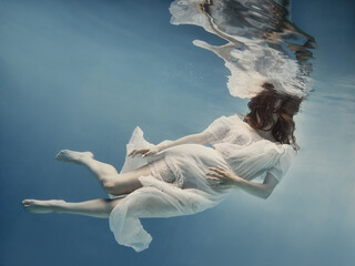 A girl with dark hair in a white dress swims underwater as if in weightlessness