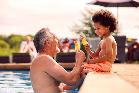 Grandfather And Grandson Eating Ice Lolly At Edge Of Swimming Pool On Summer Holiday