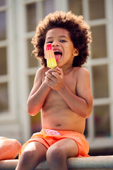 Family On Summer Holiday With Boy Eating Ice Lolly Splashing At Edge Of Swimming Pool