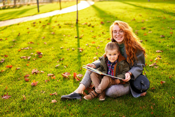 Redhead woman reads book with little boy sitting on grass in city park. Young mom teaches her first grader son and doing homework on sunny autumn day. Back to school concept. Foliage on green lawn.
