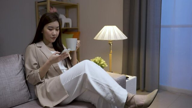 Pretty Asian Business Woman Walking To Living Room Sofa Is Sitting Down Sipping Tea And Reading Message On Mobile Phone At Leisure After Work At Home.