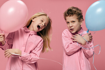  joyful, cute school-age children stand on a pink background holding pink and blue balloons in their hands, smiling joyfully and looking at each other. Horizontal studio photography with empty space