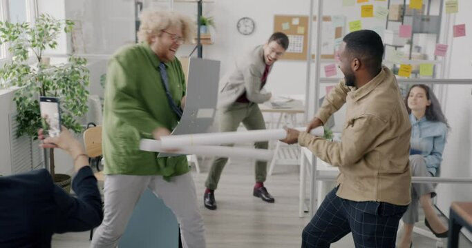 Playful men employees are fighting paper swords enjoying funny activity at work while woman colleague is recording video with smartphone
