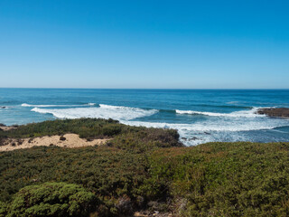 Fototapeta na wymiar View of wild Rota Vicentina coast, sea shore with ocean waves , sand and green vegetation at sunny day, clear blue sky