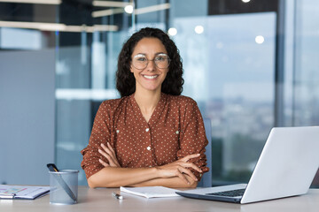 Portrait of successful young businesswoman, Hispanic woman working inside modern office building smiling and looking at camera sitting at desk with laptop, female worker with curly hair and glasses - obrazy, fototapety, plakaty