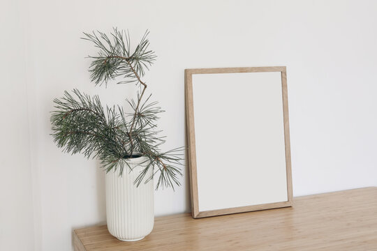 Christmas Scandinavian interior. Minimal winter artistic composition. Blank vertical wooden picture frame mockup. Pine tree branches in vase on table, desk. Beige wall background. Empty copy space.
