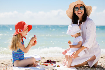 Mother and two small daughters kids sitting at sea beach picnic, eating healthy strawberries snack and smiling, happy family vacation