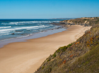 View of empty Praia do Brejo Largo beach with ocean waves, stone, wet golden sand and green vegetation at wild Rota Vicentina coast near Vila Nova de Milfontes, Portugal.
