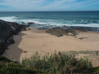 View of empty small sand beach with ocean waves and sharp rock and cllifs at wild Rota Vicentina coast near Porto Covo, Portugal.