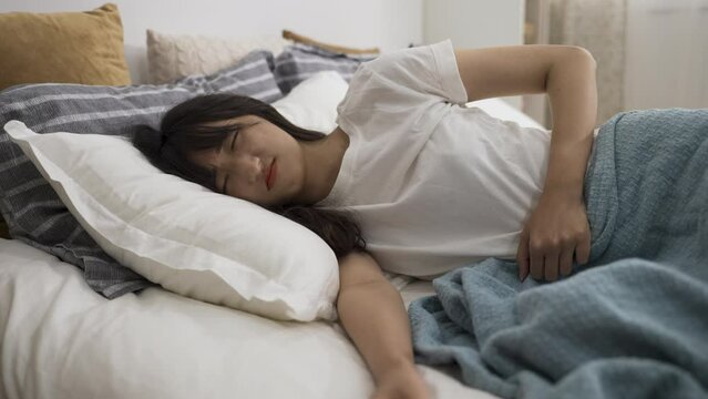 asian girl having severe cramp in lower abdomen is touching her belly and lying on bed with a painful look on face in the bedroom at home during daytime.