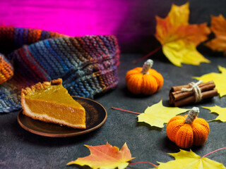piece of traditional pumpkin cake on the table with autumn yellow leaves and knitted small pumpkins. Purple backlight background