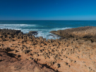 Fototapeta na wymiar View of wild Rota Vicentina coast with ocean waves and red and orange sharp rock formations near Vila Nova de Milfontes, Portugal. Sunny day, blue sky