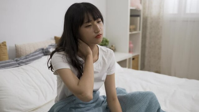 portrait of an unhappy asian girl sitting on bed and massaging her neck muscles to relieve pain in the bedroom at home.