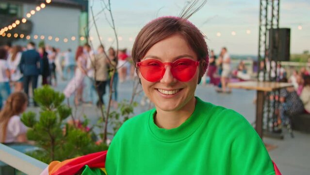 Close-up portrait of smiling young woman in trendy red glasses and green sweatshirt looking at camera in background of party in evening outdoor. Summertime. Lgbt community