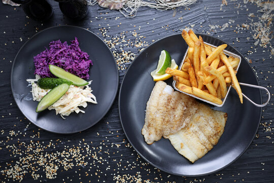 Fried fish fillets served with potato fries in a metal serving basket and salad mix, on black plates, top view.
