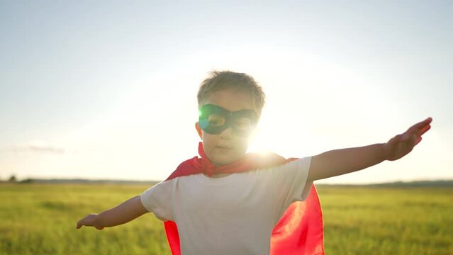 Happy child in park at sunset. Superhero winner. Boy in a red cloak and mask runs through grass.Child plays superhero in nature.Boy dressed as superhero plays in park at sunset.Childhood dream concept