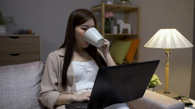 portrait asian businesswoman working overtime on computer from home is murmuring to herself in contemplation while sipping tea on sofa in the living room