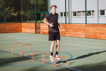 Blond boy in sportswear jumps over red hurdles to improve lower body dynamics. Plyometric training in an outdoor environment. Improve your skills