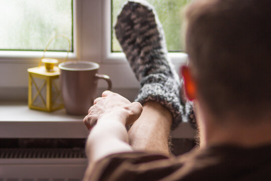Man reaches out for a cup of tea, his feet wearing in handmade knitted wool socks and lying on windowsill at cozy home.