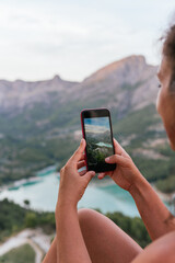 Vertical image of an unrecognizable young woman photographing with her mobile phone the mountainous landscape surrounding the turquoise blue water swamp.