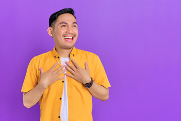 Cheerful handsome young Asian man with hands on chest and looking aside isolated on purple background - obrazy, fototapety, plakaty