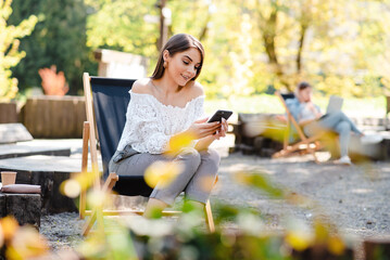 Attractive stylish smiling woman working looking smartphone, freelancer sitting in park on chair